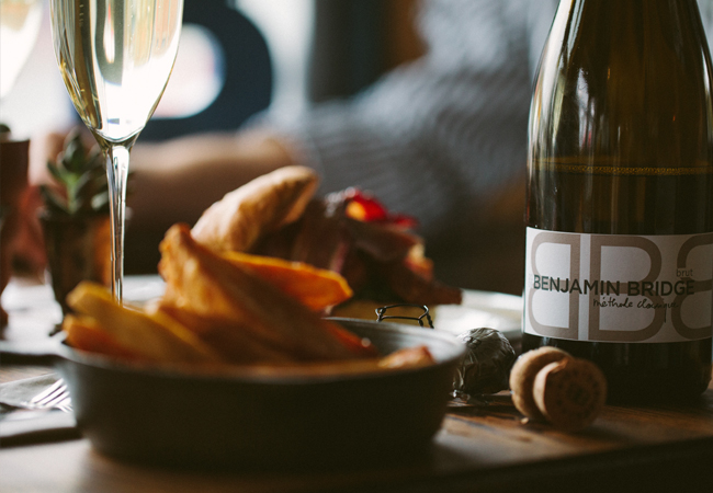 Bottle of Benjamin Bridge wine on a dining table, an appetizer in the foreground, out of focus, with a glass of white wine in the background