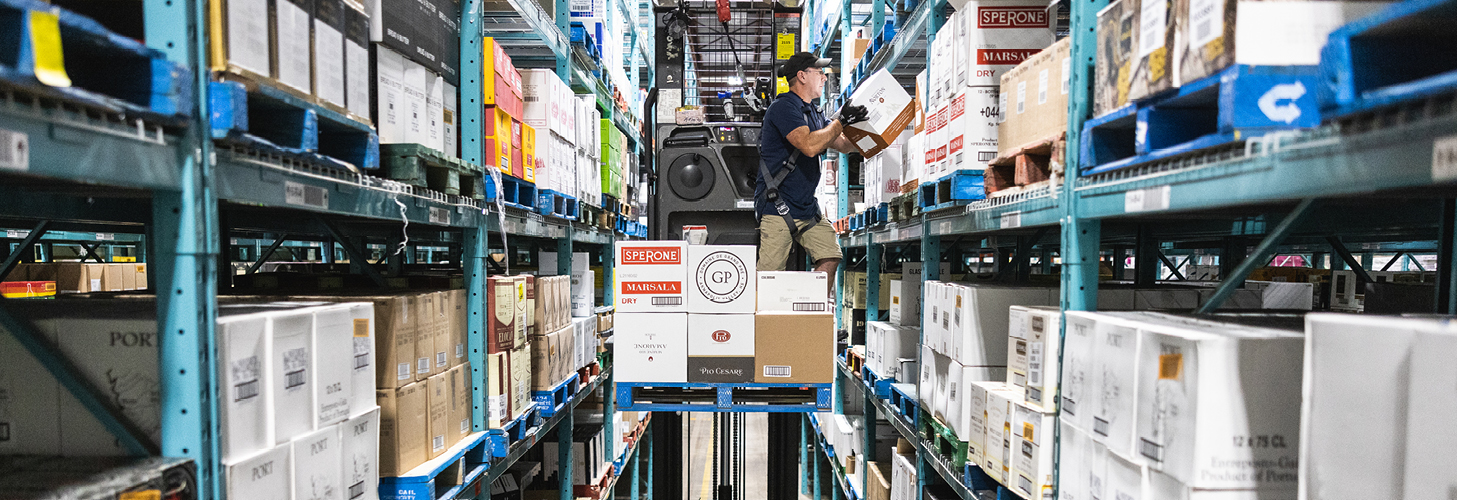 An NSLC distribution centre team member stands on a fork life with proper protective equipment stocking removing product from metal shelves.