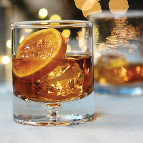 A mixed drink in a whisky glass on the rocks garnished with candied orange.