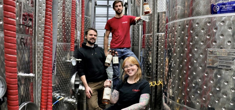 Three Compass Distillers team members sitting between large steal distilling drums 