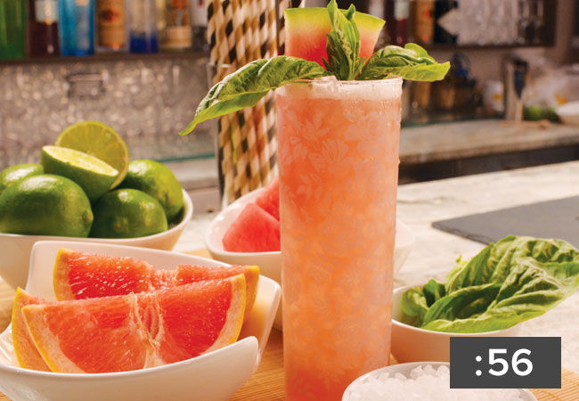 Close up of swizzle drink in a tall glass garnished with watermelon and basil