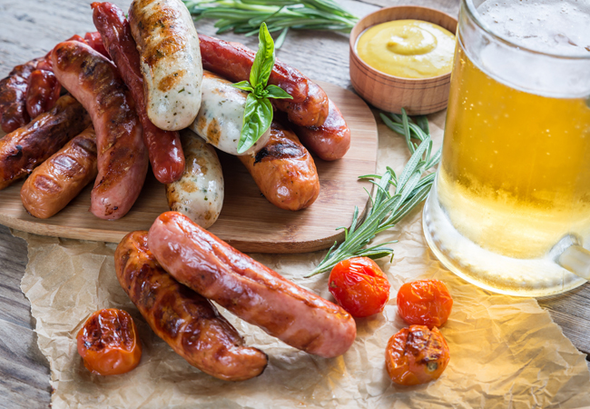 Sausages on a wooden plank beside a beer and grilled tomatoes