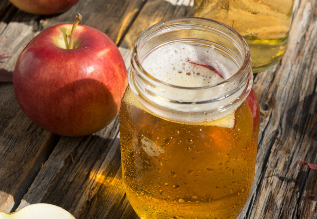 A mason jar of cider sitting on a wood plank with an apple beside it