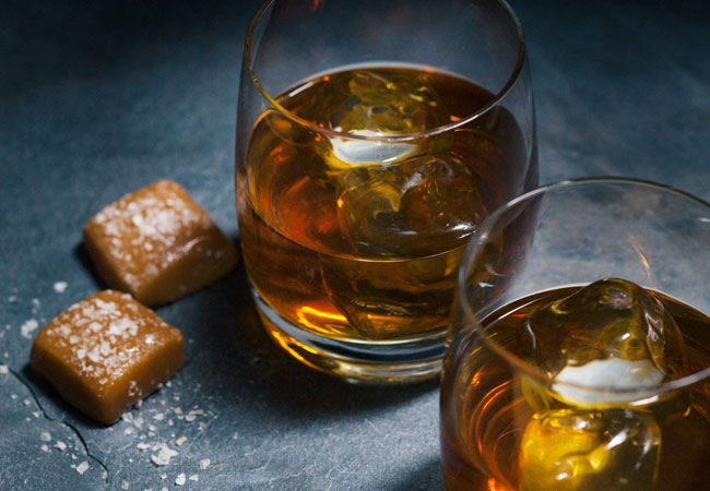 Two rocks glasses of whisky on ice with two salted caramel squares