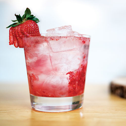 A closeup of a mostly red mixed drink in an Old Fashioned glass, served over ice and garnished with strawberry.