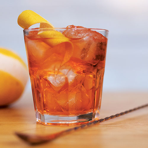 A closeup of a bright orange drink in a standard rocks glass, poured over ice and garnished with a curled grapefruit peel.