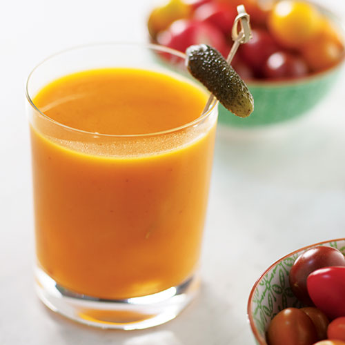 A closeup of a caesar made with a yellow tomatoes purée that is garnished with a Gherkin pickle on a stick.