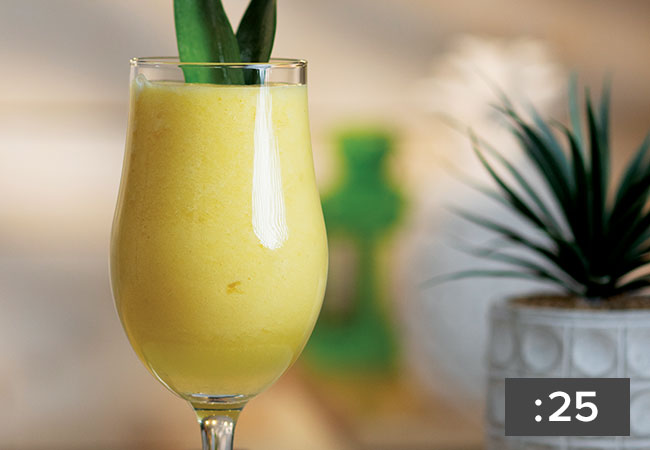 A closeup of a yellow smoothie in a poco grande glass garnished with the leaves from a pineapple. Recipe video is 25 seconds.