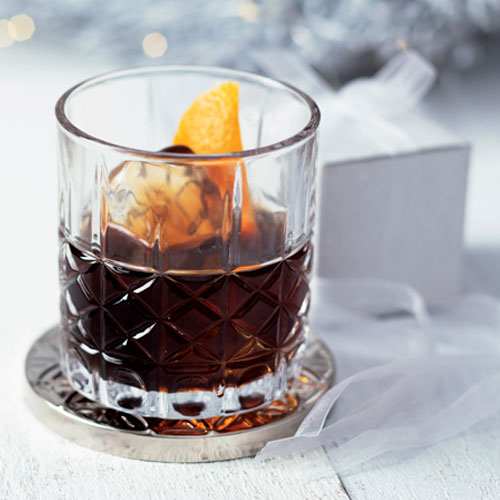 A drink in a faceted old fashioned glass on the rocks with a lemon peel in it, sitting on a coaster in front of a present with a bow.