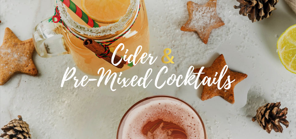 A top view of mixed holiday drink in a mason jar on a table that is decorated with star-shaped cookies, pinecones and fake snow. Text that says “Cider & Pre-Mixed Cocktails”.