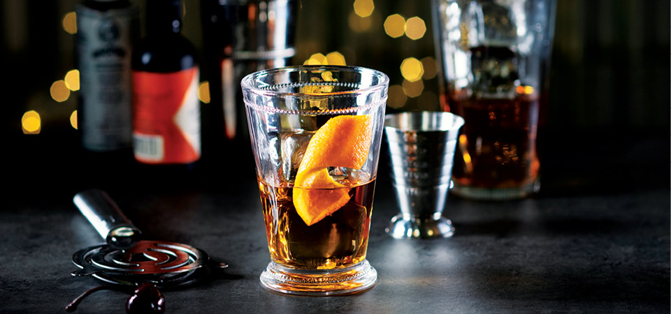 A detailed highball glass containing a transparent light brown whisky and a curled slice of orange peel.