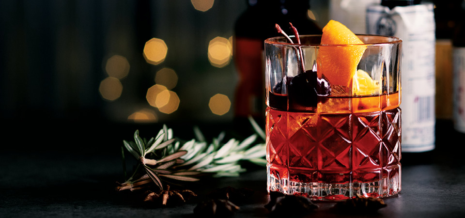 A detailed old fashioned glass containing a transparent reddish brown whisky topped with black cherries and an orange peel.