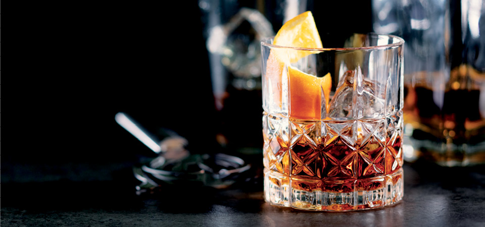An ornate old fashioned glass containing transparent light brown whisky topped with a curled orange peel and square ice cubes.