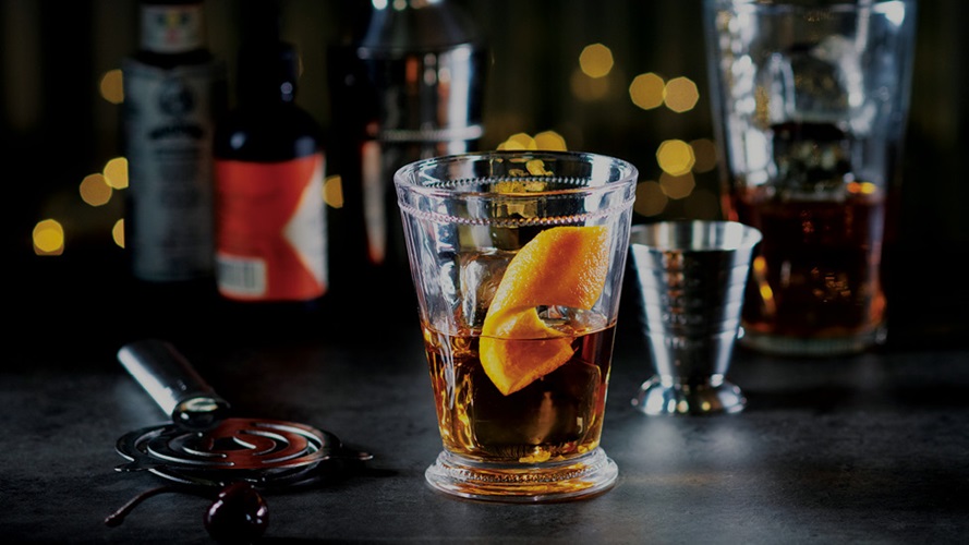 A detailed highball glass containing a transparent light brown whisky and a curled slice of orange peel.
