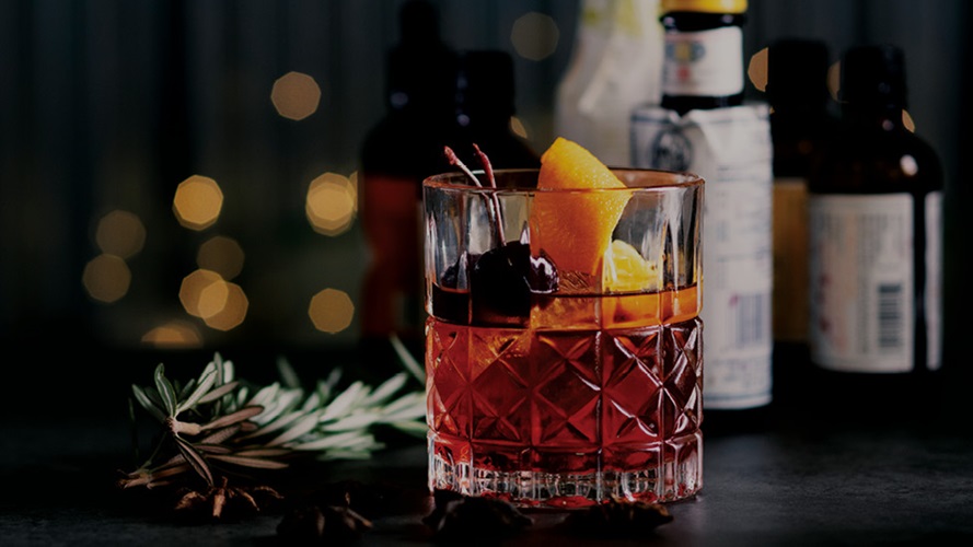 A detailed old fashioned glass containing a transparent reddish brown whisky topped with black cherries and an orange peel.