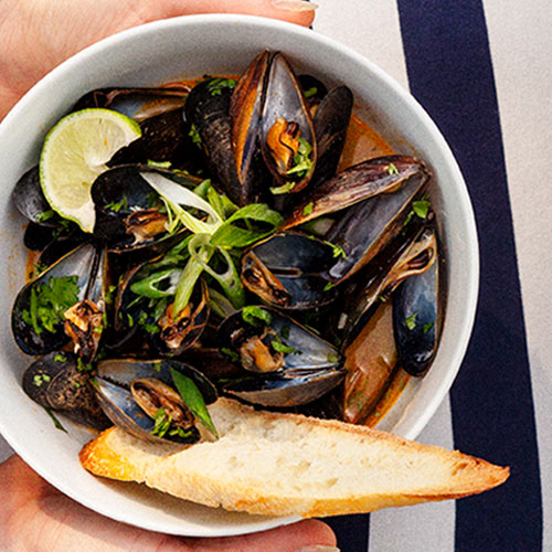 A bowl of mussels in a red broth served with a piece of baguette