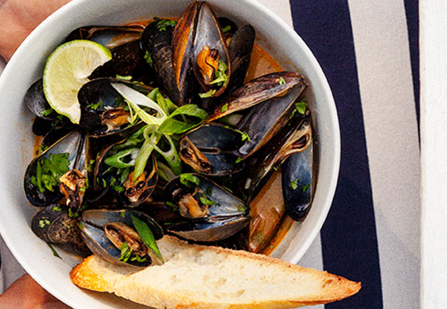 A bowl of mussels in a red broth served with a piece of baguette