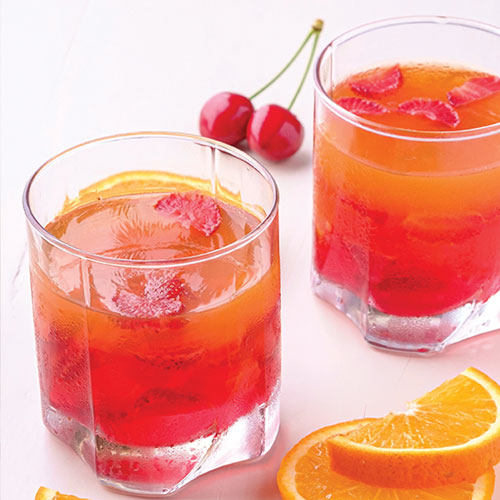 Two orange-red cocktails with cherries in them, surrounded by a cherry and orange slices.