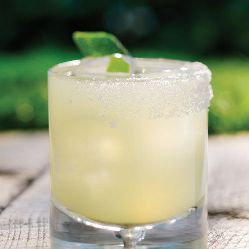 Margarita in a short glass with salted rim and lime wedge sitting on a wooden table outside.