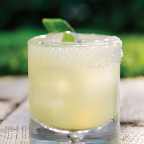 Margarita in a short glass with salted rim and lime wedge sitting on a wooden table outside. 