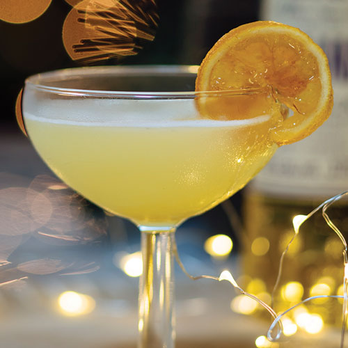 Light yellow cocktail with a golden lemon slice for garnish and twinkling lights in the background. 