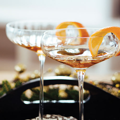 On a black tray sits two cocktails in crystal wine glasses with an orange rind for garnish. 