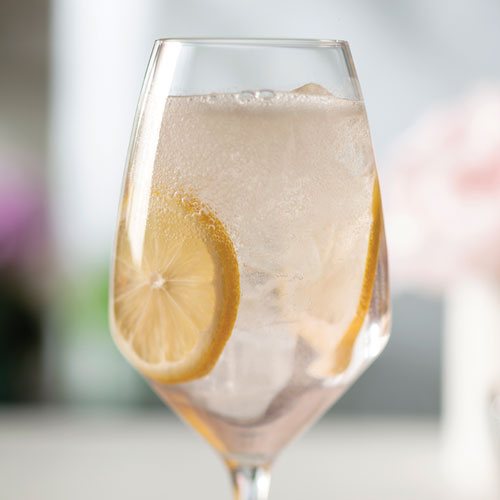Fizzing, light pink cocktail in a wine glass with ice cubes and lemon slices.