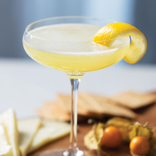 A yellow cocktail in a martini glass garnished with a lemon peel.