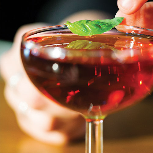 A deep red cocktail with someone placing a basil leaf on top. 