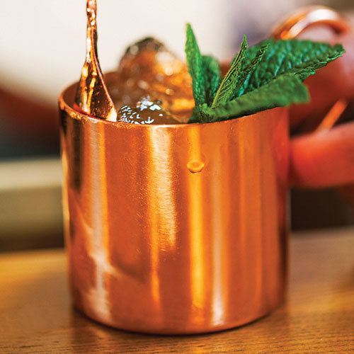 Splashing liquid into a copper mug filled with ice, topped with mint garnish.