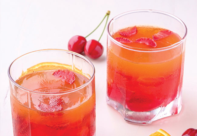 Two orange-red cocktails with cherries in them, surrounded by a cherry and orange slices.