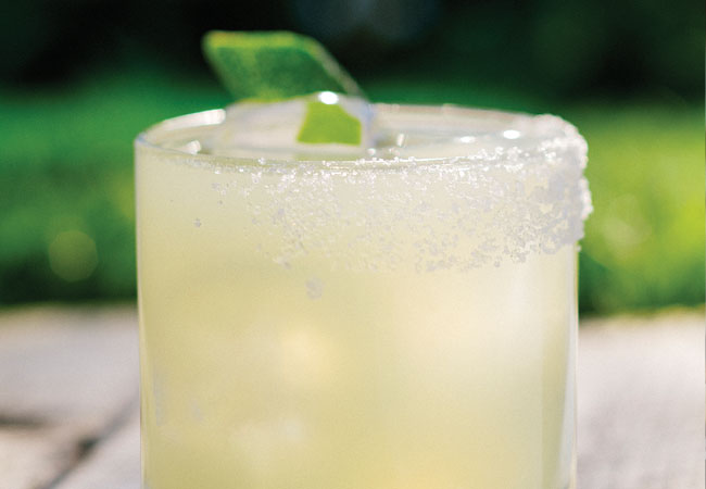 Margarita in a short glass with salted rim and lime wedge sitting on a wooden table outside. 