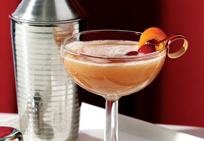 Cocktail with cranberry and orange peel garnish sitting on a white tray with a shaker in the background. 
