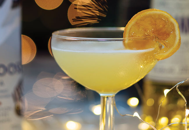 Light yellow cocktail with a golden lemon slice for garnish and twinkling lights in the background. 