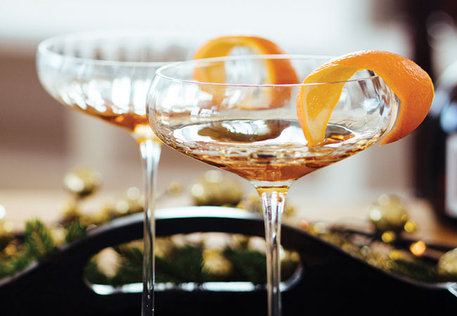 On a black tray sits two cocktails in crystal wine glasses with an orange rind for garnish. 