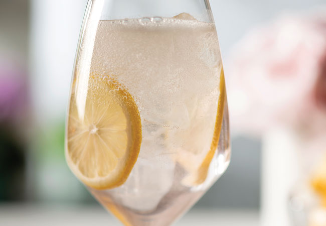 Fizzing, light pink cocktail in a wine glass with ice cubes and lemon slices. 