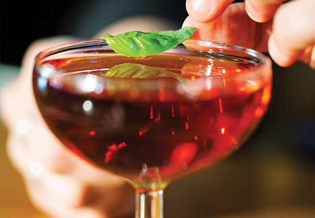 A deep red cocktail with someone placing a basil leaf on top. 