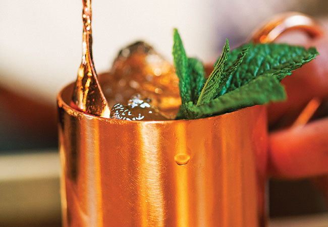 Splashing liquid into a copper mug filled with ice, topped with mint garnish.