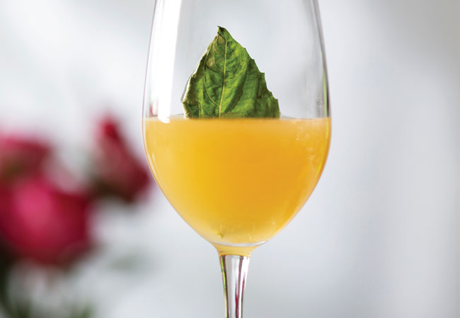 Bright yellow cocktail in a wine glass with a basil leaf garnish. 
