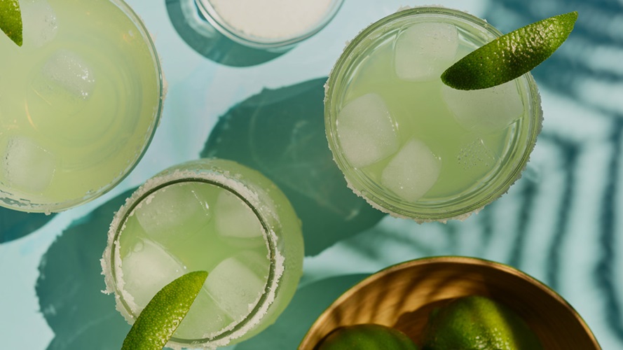 Three pale green drinks in various salt-rimmed glasses with a lime wedges.