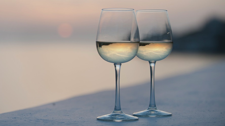 A pink and blue sunset in the background of the shot tints two wine glasses filled with a light white wine.