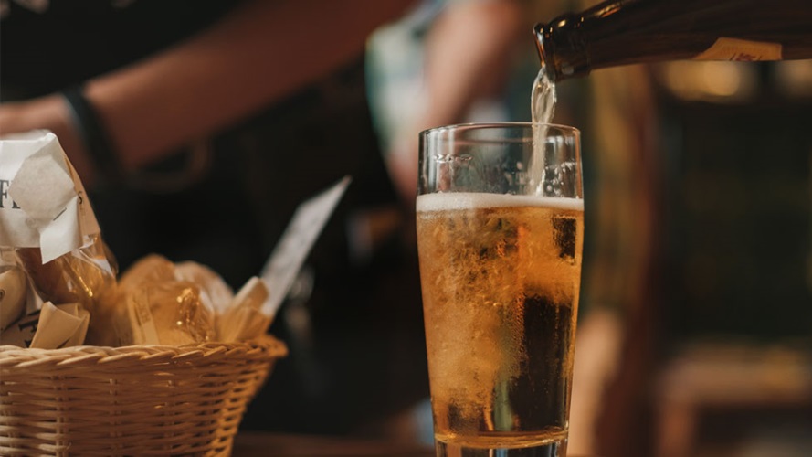 A bottle of beer is being poured into a glass with a basket of bread in the background.