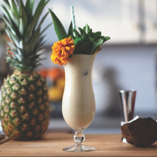Tropical creamy white cocktail garnished with an orange flower and green foliage 