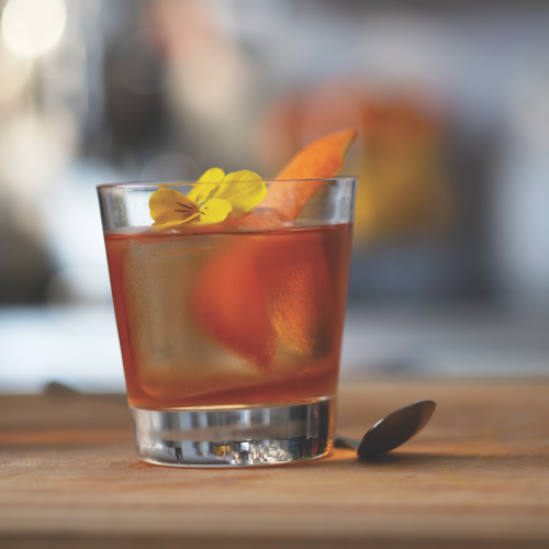 Cocktail glass filled with an orange liquid garnished with a yellow flower and orange peel