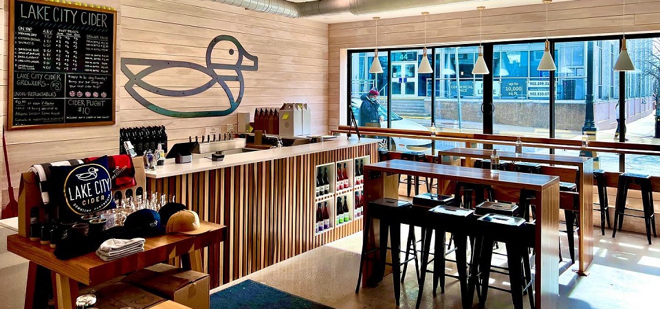 View inside of Lake City Ciders Tap room. Hightop tables in the middle of the room with a counter with a large image of a duck. 
