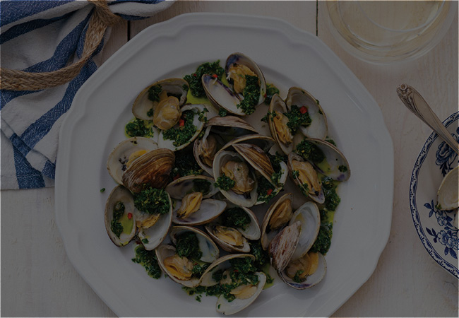 A white bowl full of clams with a salsa verde sits on a table with a blue and white napkin off to the side