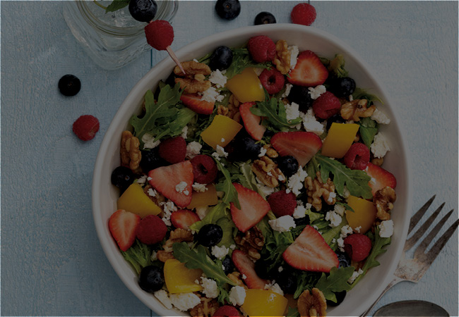 A white bowl at a table setting filled with a salad with berries, oranges and feta cheese 