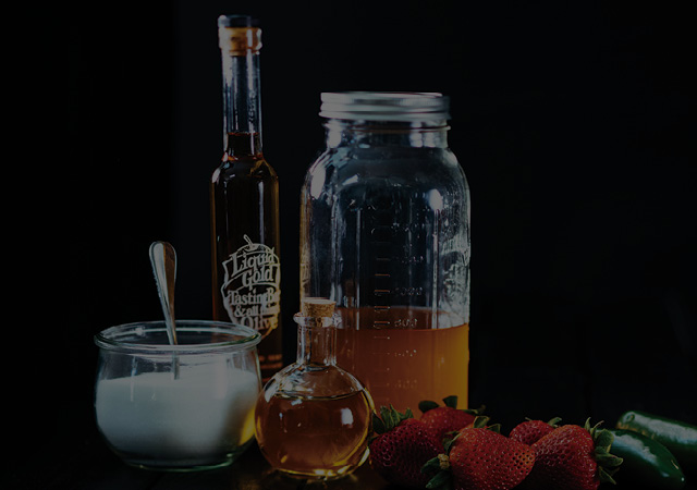 Sugar, apple cider vinegar, strawberries, jalapenos and balsamic vingar sit on a table with a black background, with a large mason jar filled half way with the Strawberry-jalapeno shrub.  