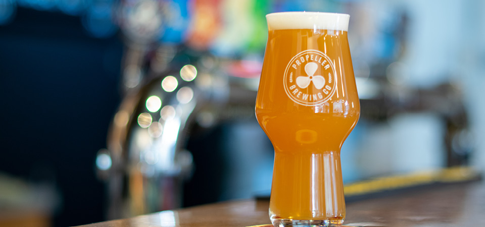 A glass of light coloured Propeller beer sits on a bar with taps in the background.