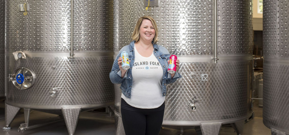 Woman standing in front of cider brewing equipment holding two different cans of Island Folk cider.